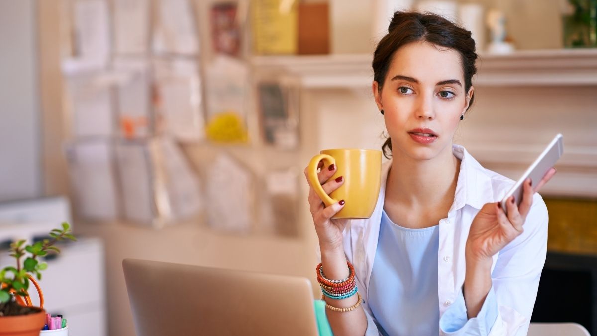 Young woman holding a phone and coffee cup while working at her desk, contemplating how much should I contribute to my HSA.