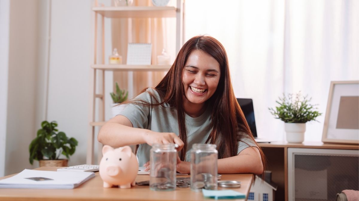 Smiling woman saving money in jars at home with a piggy bank on the table, representing the comparison between an HSA vs FSA account for healthcare savings.